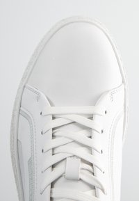Zapatilla blanca con parte superior de cuero suave, puntera texturizada, cordones planos y detalles de costura sutiles. Suela de goma y ligero relieve decorativo.