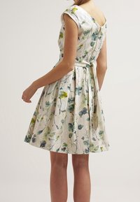 Kukallinen mekko lyhyillä hihoilla, V-mallisella selällä ja laskostetulla hameella. Kankaan kuvioissa on vihreitä ja keltaisia kukkia vaalealla taustalla.