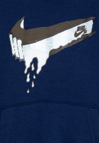 Sötétkék pulóver, amelyen egy nagyméretű, csöpögő fehér Nike logó található szürke részletekkel, középen a mellkason. Puha anyagtextúra látható.