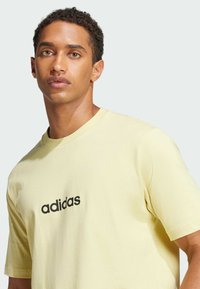 Žluté bavlněné tričko s krátkým rukávem, na přední straně se středovým černým logem "adidas", kulatým výstřihem a volným střihem.