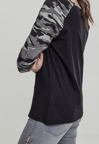 Person som bär en svart skjorta med grå kamouflage långärmar och ljusgrå jeans med snörningsdetalj på låret, poserar från sidan.