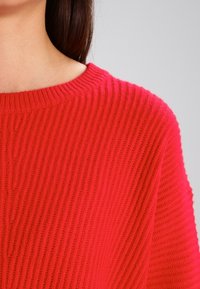 Red knitted sweater featuring a ribbed texture and a wide crew neckline. The fabric appears soft and cozy with a chunky stitch design.