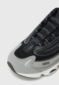 Sneaker noir et gris avec une tige en maille, une languette rembourrée et une semelle en caoutchouc avec des coutures visibles et un détail "1000" sur le côté.