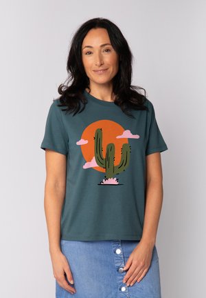 Teal katoenen t-shirt met een cactusafbeelding, een oranje cirkel en roze wolken. Klassieke pasvorm met korte mouwen, gladde textuur.