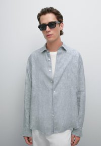 Young man wearing rectangular black sunglasses, loose gray striped button-up shirt over white top, and white pants against a plain background.