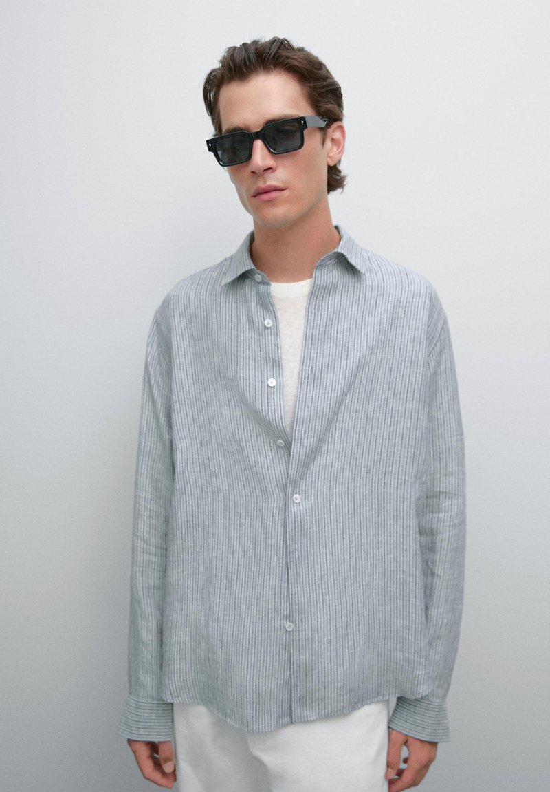 Jeune homme portant des lunettes de soleil noires rectangulaires, une chemise à rayures grises lâche par-dessus un haut blanc, et un pantalon blanc, se tenant devant un fond uni.