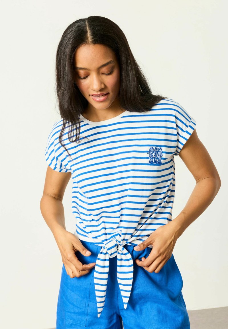 Frau richtet ihr gebundenes blau-weiß gestreiftes Hemd, trägt leuchtend blaue Shorts und steht vor einem einfarbigen hellen Hintergrund.