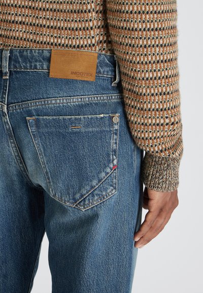 El bolsillo trasero de unos jeans de mezclilla azul con una etiqueta de cuero marrón de la marca Incotex, combinado con la manga de un suéter de punto de tonos marrones y beige.