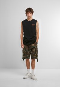 Schwarzes ärmelloses Shirt mit orangefarbenem Text, kombiniert mit camouflaged Cargo-Shorts. Weiße Sneakers mit einem strukturierten Design und knöchellangen weißen Socken.