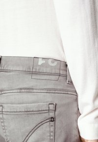 Grå denimjeans med en slät textur, med en konturerad bakficka och en etikett som visar siffran "24." En vit skjorta är delvis synlig.