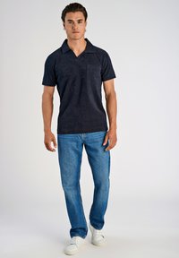 Navyblauw polo shirt met een borstzak en korte mouwen, gecombineerd met lichtblauwe jeans en witte sneakers, staande tegen een effen achtergrond.