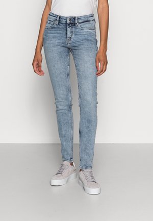 Slim-fit hellblaue Jeans mit Used-Waschung, getragen mit weißen Sneakers und einem weißen, in die Hose gesteckten Top, stehend vor einem schlichten hellen Hintergrund.