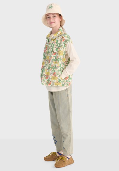 Młoda osoba ubrana w kamizelkę w kwiatowy wzór, beżowy kapelusz typu bucket, jasną koszulę z długim rękawem, luźne dżinsy z niebieskimi detalami oraz brązowe mokasyny.