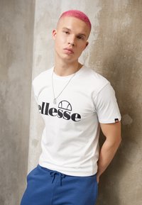 Hvit T-skjorte med svart logo, tettsittende design, kombinert med blå joggebukser. Modellen har kort rosa hår og et sølvkjede med et anheng.