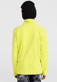 Pull en polaire jaune avec un col haut, une coupe ajustée et des manches longues. Porté avec un bonnet noir et un pantalon gris foncé à motifs.