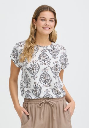 Lachende vrouw met lang blond haar die een wit, patroonig shirt met korte mouwen en beige broek draagt, met haar handen in de zakken.