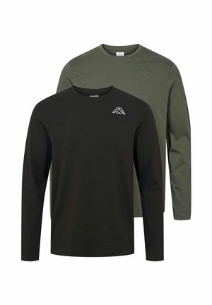 Férfi áll, kezét a zsebében tartva, fekete hosszú ujjú inget, fekete oldalcsíkos tréningnadrágot és fehér tornacipőt visel, egyszínű háttéren.