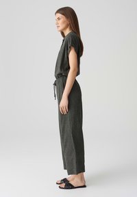 Dunkelgrüner Jumpsuit mit kurzen Ärmeln, taillierter Taille und lockerer Passform. Hergestellt aus strukturiertem Stoff mit subtilem Muster. Kombiniert mit schwarzen Sandalen.