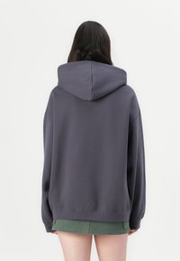 Overdimensjonert mørkegrå hettegenser med en romslig hette og lange ermer, laget av mykt stoff, kombinert med et grønt skjørt. Glatt tekstur, minimal detaljering.