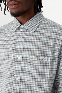 Homme portant une chemise à carreaux bleu clair et marron à boutons, avec col et poche poitrine, visible du cou jusqu'au milieu du torse.