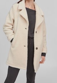 Abrigo de forro polar esponjoso de color crema con cuello con muescas, dos bolsillos laterales y dos botones en la parte delantera. Llevado sobre una parte superior y un pantalón gris oscuro.