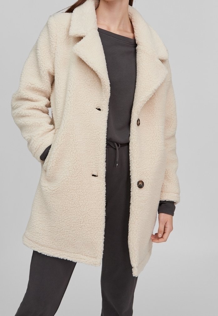 Abrigo de forro polar esponjoso de color crema con cuello con muescas, dos bolsillos laterales y dos botones en la parte delantera. Llevado sobre una parte superior y un pantalón gris oscuro.