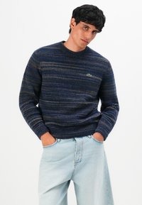 Uomo che indossa un maglione a righe blu navy e jeans azzurri, con le mani nelle tasche, in piedi davanti a uno sfondo bianco semplice.