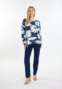 Donkerblauwe trui met grote witte bloemmotieven, gecombineerd met donkerblauwe broek en lichtbruine hakken. Zachte textuur, ontspannen snit en lange mouwen.