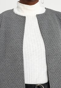 Lähikuva henkilöstä, joka on pukeutunut teksturoituun harmaaseen avonaisfronttiseen takkiin valkoisen ribbivillapaitasvein ja mustan vyön, jossa on hopea solki.