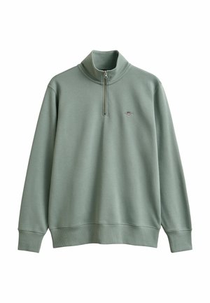 Lichtgroene pullover met lange mouwen, halve ritskraag en klein geborduurd schildlogo op de linkerborst.