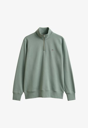 Lichtgroene pullover met lange mouwen, halve ritskraag en klein geborduurd schildlogo op de linkerborst.