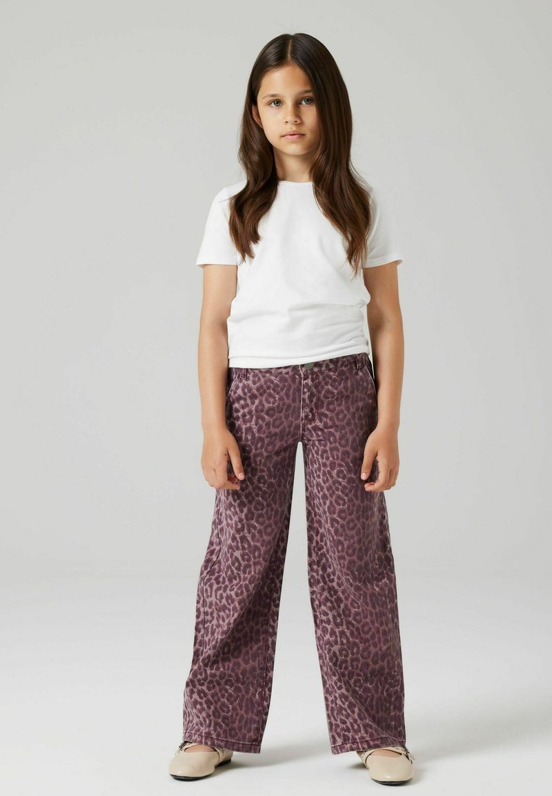 Meitene ar gariem brūniem matiem stāv, ģērbusies baltā T-kreklā, platās violetās leopardu raksta biksēs un beiges kurpes uz neitrāla fona.