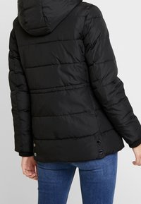 Chaqueta acolchada negra con capucha, con un diseño acolchado, puños de canalé y detalles de botones a presión en el dobladillo. Textura suave.