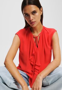 Blusa sin mangas de color rojo con escote tipo ojal y detalle de volantito en el frente, hecha de tela ligera. La modelo lleva jeans de color azul claro.