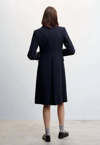 Cappotto di lana blu scuro con una forma strutturata, maniche lunghe e spacco sul retro. Indossato sopra calze grigie e scarpe scure, visto da dietro.