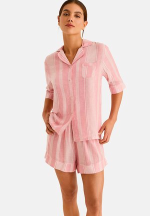 Femme portant une chemise à boutons rayée verticale rose et blanche avec un short ample assorti, sur un fond uni.
