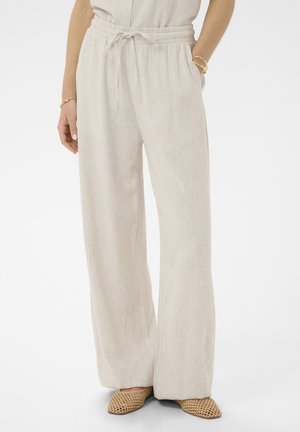 Nő világos bézs színű, bőszárú nadrágot visel, amelynek húzózsinóros derékrésze van, fonott, barna slip-on cipőt és arany karpereceket a csuklóin.