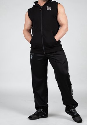 Persona muscolosa che indossa una felpa nera senza maniche con zip, pantaloni neri larghi con logo e scarpe da ginnastica alte nere, in piedi con le mani in tasca.