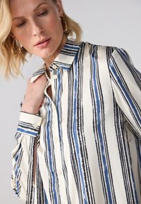 Blusa a righe color crema, con linee blu e nere. Design con bottoni e colletto, maniche lunghe. Tessuto liscio e setoso.