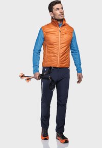 Herren-Outfit für den Outdoor-Bereich mit einer orangefarbenen Steppweste, einem blauen Langarmhemd, einer navyblauen Hose und schwarzen-orangefarbenen Wanderschuhen. Enthält Trekkingstöcke.