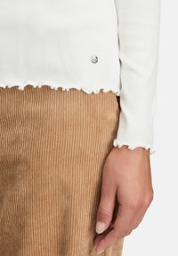 Weißes geripptes Langarmoberteil mit fransigem Saum, kombiniert mit einer strukturierten braunen Cordhose. Dezente metallische Akzentuierung am Saum.