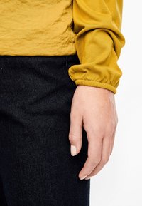 Långärmad, strukturerad skjorta i senapsgul med sammanknutna manschetter, tillsammans med mörka denimjeans. En hand vilar mot sidan, omärkt.