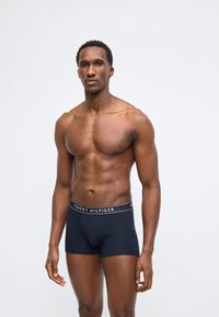 Μυώδης άνδρας που στέκεται με το πρόσωπο προς τα εμπρός, φοράει σκούρο μπλε μποξέρ Tommy Hilfiger, ουδέτερη έκφραση, απλό ανοιχτό φόντο.