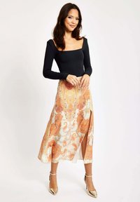 Black long-sleeve top with square neckline, paired with a patterned orange and cream skirt featuring a side slit. Gold high-heeled shoes.