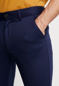 Námořnické modré kalhoty na míru s hladkým povrchem, se přední kapsou a zapínáním na knoflík. V doprovodu hořčicové košile.