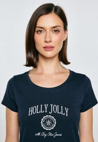 Tumši zils t-krekls ar īsām piedurknēm, apaļu kakla izgriezumu un baltiem izdrukātiem vārdiem "HOLLY JOLLY" ar apļveida dizainu zemāk. Mīksta auduma tekstūra.
