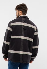 Černá kostkovaná bunda se zelenými a béžovými pruhy, vyrobená z texturované tkaniny. Má límec a manžety na knoflíky, zobrazeno zezadu.