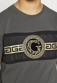 Šedé bavlněné tričko s černým pruhem a zlatou vyšitou logem a geometrickými vzory na hrudi.