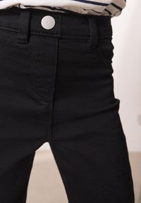 Jeans de mezclilla negra con un diseño ajustado, que cuentan con un cierre de botón metálico redondo, bolsillos frontales y detalles de costura sutiles.