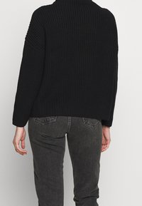 Czarny sweter wykonany na drutach z wysokim dekoltem, luźnym kroju i prążkowaną fakturą, w zestawieniu z ciemnoszarymi dżinsami z tylnymi kieszeniami.